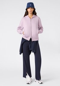 Felpa zip-up lilla, abbinata a pantaloni larghi blu navy e un cappello a secchiello in denim. Presenta tasche frontali e sneakers bianche.