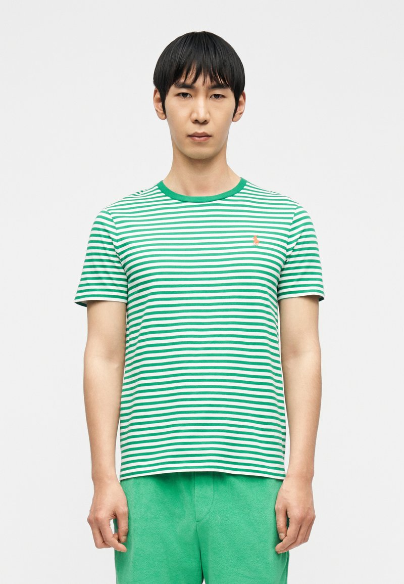 Άνδρας με κοντά μαύρα μαλλιά που φοράει πράσινο  και λευκό ριγέ T-shirt και πράσινο παντελόνι, στέκεται μπροστά σε απλό ανοιχτό φόντο.