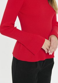Top lungo a maniche con righe rosse e polsini svasati, caratterizzato da un design aderente e una morbida texture, abbinato a pantaloni neri.