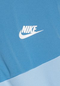 Zila Nike auduma ar gludu tekstūru, kurā izceltas baltas krāsas Swoosh logo. Izteikta krāsu bloku dizaina iezīme ar gaišāku zilu apakšā.