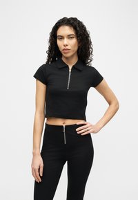 Γυναίκα με σγουρά μαλλιά που φοράει μαύρο κοντό πόλο με φερμουάρ και μαύρο παντελόνι ψηλόμεσο με φερμουάρ στο μπροστινό μέρος, στέκεται με το ένα χέρι στη μέση.