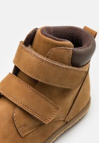 Braune Wildleder-Stiefelette mit zwei Klettverschlüssen, einem gepolsterten braunen Innenraum und einer verstärkten braunen Ferse. Langlebige Nähte und Gummisohle.