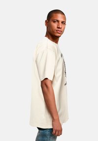 Beige Baumwoll-T-Shirt mit kurzen Ärmeln, lockerer Passform und Graf printed. Getragen mit blauen Jeans, von der Seite gezeigt.