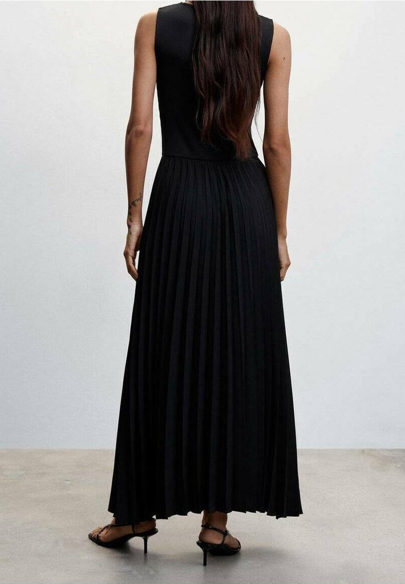 Femme aux longs cheveux portant une robe longue noire plissée sans manches et des sandales à talons noires à brides, debout contre un mur uni.