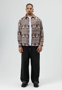 Chemise marron à motifs avec des dessins de cerfs, boutonnée, portée sur un t-shirt blanc, associée à un pantalon noir ample et des chaussures noires.