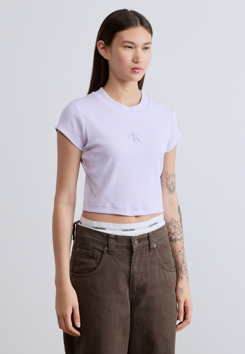 Λιλά cropped t-shirt από ριμπ ύφασμα με ρεβέρ μανίκια και κεντημένο λογότυπο «CK». Συνδυάζεται με καφέ παντελόνια ψηλόμεσα που διαθέτουν εμφανή λωρίδα με λογότυπο.