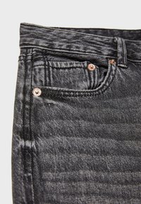 Gros plan sur un jean en denim noir montrant la poche avant, la poche à monnaie, le passant de ceinture et les rivets en cuivre avec texture de tissu et coutures visibles.