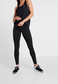 Débardeur de sport noir à coupe ample, associé à un legging noir et des baskets noires avec des accents blancs. Design minimaliste, texture lisse.