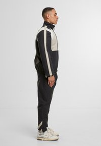 Completo giacca e pantaloni in tonalità nero e beige. Presenta un colletto alto, chiusura con zip e un tessuto morbido con una finitura leggermente strutturata.