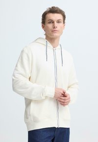 Sweat à capuche zippé beige clair avec un tissu texturé, des cordons de serrage gris et une fermeture éclair bleue contrastante. Comprend une capuche et des poignets côtelés.
