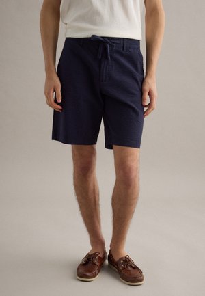 Mann trägt marineblaue, strukturierte Shorts mit Kordelzug, ein weißes ärmelloses Oberteil und braune Leder-Bootsschuhe, steht vor einem neutralen Hintergrund.