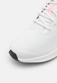 Zapatilla deportiva blanca con parte superior de malla texturizada, que presenta acentos rosas en la marca y una suela de goma negra para mayor tracción.