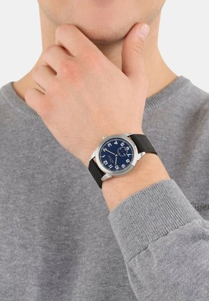 Analoge Uhr mit blauem Zifferblatt, silbernem Gehäuse und schwarzem Lederarmband. Mit großen weißen Stunden-Ziffern und einem Datumfenster.