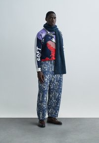 Ανδρικό σύνολο με πολύχρωμο πουλόβερ με το κείμενο «SOFT». Συνδυασμένο με μπλε παντελόνι με σχέδιο και σκουρόχρωμο κασκόλ. Καφέ παπούτσια.