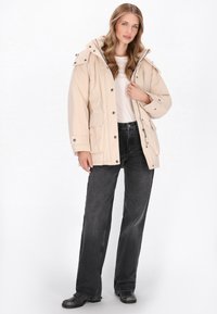 Beige waterdichte parka met capuchon, voorrits, drukknoopsluiting en zijzakken. Gecombineerd met zwarte wijde jeans en zwarte schoenen.