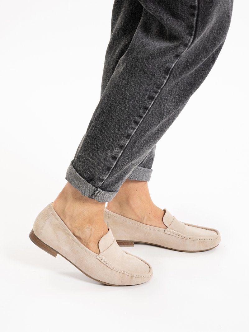 Person iført op rullede mørkegrå jeans og lysebeige loafers i ruskind med træsåler på en hvid baggrund.
