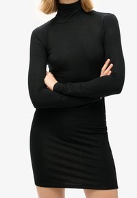 Frau in einem taillierten schwarzen Langarmrollkragenkleid mit verschränkten Armen vor einem schlichten weißen Hintergrund.