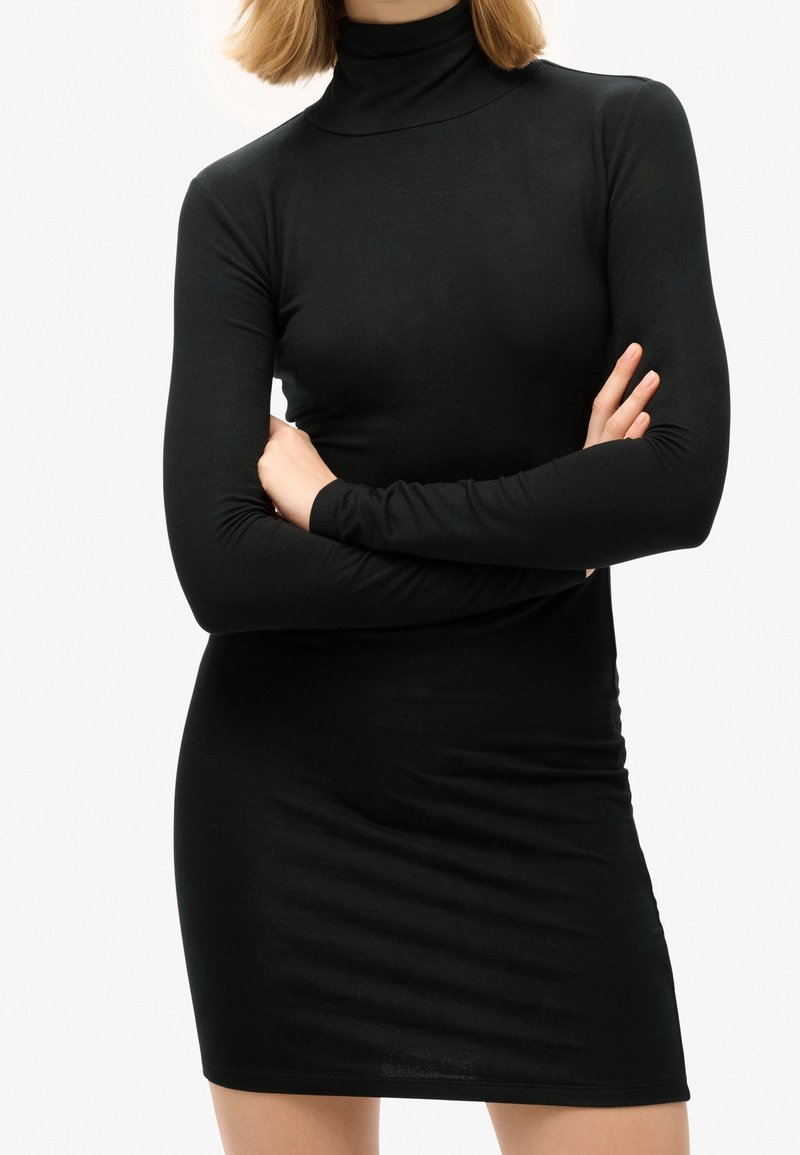 Frau in einem taillierten schwarzen Langarmrollkragenkleid mit verschränkten Armen vor einem schlichten weißen Hintergrund.