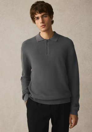 Grå strikket pullover med halvlås krave, struktureret stof og ribbede manchetter. Parret med sorte bukser. Casual, struktureret design.