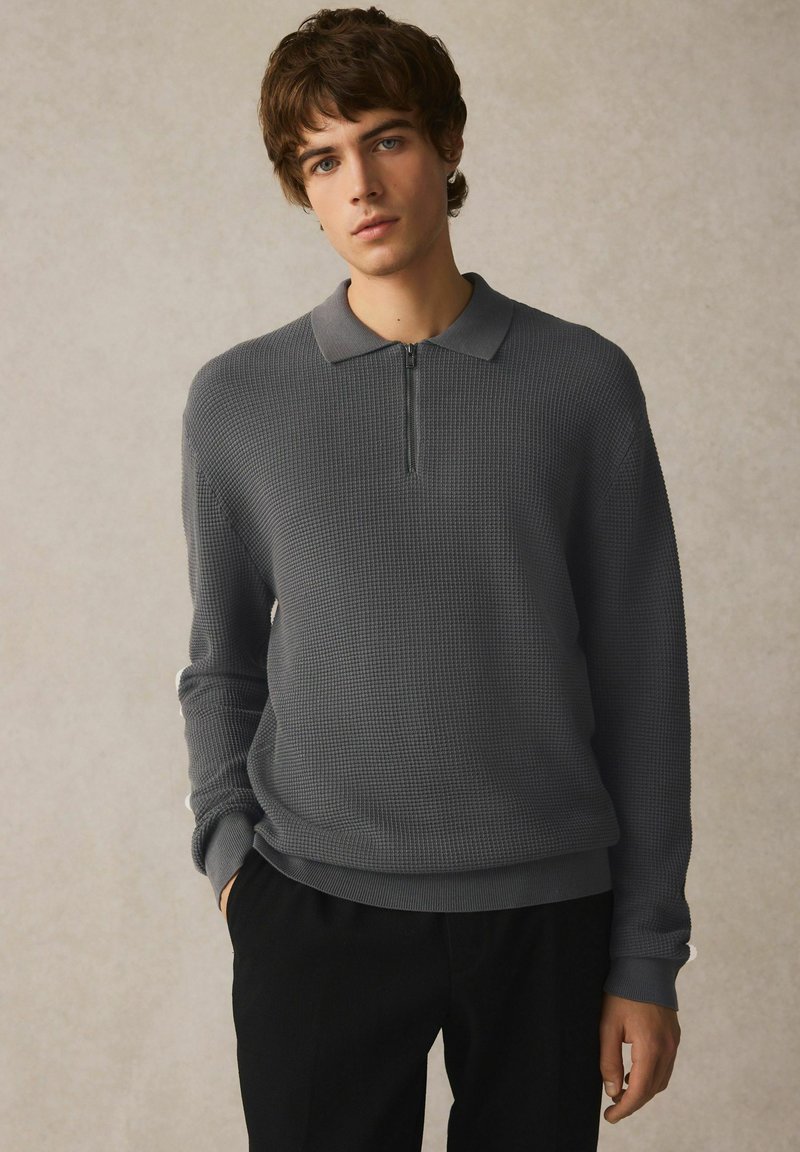Grå strikket pullover med halvlås krave, struktureret stof og ribbede manchetter. Parret med sorte bukser. Casual, struktureret design.