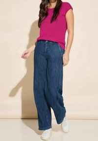 Fuchsiová krátkosrstá halenka spárovaná s volně střiženými tmavými džínami, které mají rovný střih, ohrnutý lem a bílé tenisky.