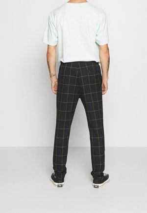 Zwarte geruite broek met een slimfit ontwerp, voorzien van gele lijnen in het raster, een enkele achterzak, en gecombineerd met een lichtgroene t-shirt en sneakers.