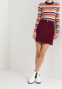 Top de manga larga a rayas con cuello alto en tonos rojos, amarillos y azules. Falda A de pana burdeos. Botines blancos brillantes con acentos negros.