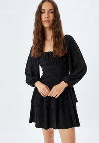 Musta mekko, jossa on pitkät puff-hihat, tiukka body, kerroksellinen röyhelöhame ja koko matkalla oleva tekstuurinen kukkakuosi.