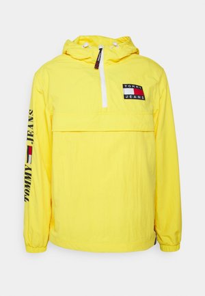 Gul huva jacka i lätt material, med halvdragkedja fram, framficka och logotypdetaljer på bröstet och ärmen.