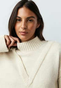 Kremfarvet strikket sweater med ribbet høj krave og V-formet udskæring. Har tekstureret stof og afslappet pasform.