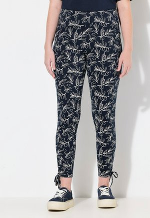 Leggings a fantasia navy con design botanico bianco; forma aderente, legacci alla caviglia, abbinati a sneakers navy a bassa altezza su uno sfondo chiaro.