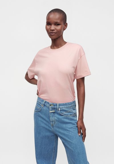 Personne debout, main sur la hanche, portant un t-shirt rose pâle à manches courtes rentré dans un jean bleu taille haute, sur un fond uni.