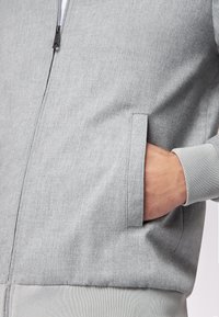 Giacca grigia con zip e materiale testurizzato, dotata di una tasca laterale e polsini a coste. Vestibilità slim con un design semplice e pulito.