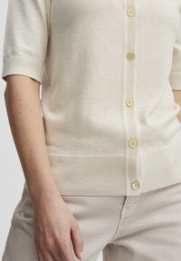 Lys beige strikket cardigan med korte ærmer, med ribbede kanter og fem dekorative gyldne knapper foran.