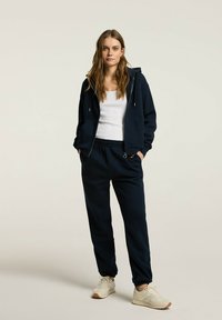Granatowy hoodie z regulowanym sznurkiem i kieszeniami z przodu, w połączeniu z dopasowanymi joggingami. Luźna biały top na ramiączkach i jasne sneakersy.
