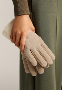 Bež zamšaste rokavice s štirimi prsti in odprtim manšetnim delom, ki jih drži roka, ob mehkem belem tekstilu, na zeleni podlagi.