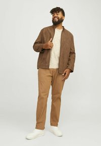 Chaqueta de ante marrón con un cuello alto, superpuesta sobre una camisa beige clara. Combinada con pantalones de color canela y zapatillas blancas, de pie de manera casual.