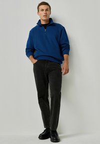 Sudadera de forro polar azul con cremallera de cuarto y mangas largas, combinada con jeans negros y zapatos negros. Diseño simple, textura suave.