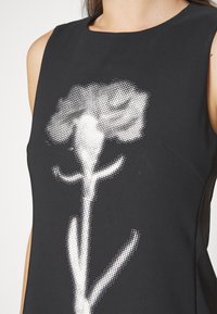 Haut noir sans manches avec un grand motif de rose blanche pixellisée à l'avant, porté par une personne montrant le haut du torse.