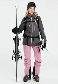 Skiausrüstung mit einer grauen Jacke mit pinken Akzenten, schwarzen Hosen und schwarzen Skischuhen. Hält Skier und Stöcke, trägt eine Strickmütze und eine Skibrille.