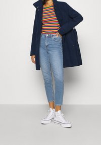 Marinblå kappa med hög krage, randig skjorta i rött, grönt, gult och blått, ljusblå smala jeans och vita högskor.