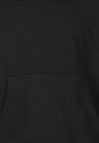 Lähikuva mustast sweatshirtist, mille eesmisel taskul on peenelt tikitud logo rinna lähedal, tumedal kangasel taustal.