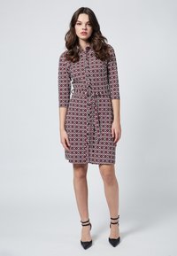 Punainen ja valkoinen kuvioitu mekko, jossa on napit, kolme neljännesosaa hihasta, vyö ja suora leikkaus, yhdistettynä mustiin kärjellisiin korkokenkiin.