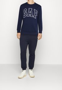 Sweat-shirt bleu marine avec logo "GAP" blanc sur la poitrine, assorti à un pantalon de jogging et des baskets blanches, avec une coupe décontractée.