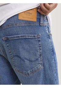 Jean en denim bleu avec une finition texturée, dotés d'une patch en cuir à la taille, de détails sur les poches arrière et de surpiqures orange.