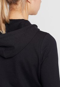 Sudadera negra con una textura suave y lisa. Cuenta con capucha con cordón y mangas raglán. La tela tiene un ligero estiramiento.