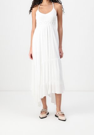 Kobieta w białej, bezrękawnej sukience maxi z koronkowym gorsetem i asymetrycznym dołem, w zestawieniu z beżowymi, spiczastymi balerinami z paskami wokół kostek.