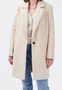 Beige uldfrakke med slået krave, enkelt knaplukning, sidelommer og lange ærmer, båret over en lys sweater og jeans.