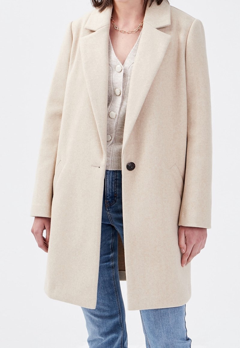 Beige uldfrakke med slået krave, enkelt knaplukning, sidelommer og lange ærmer, båret over en lys sweater og jeans.