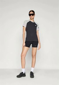 Donna che indossa un completo sportivo nero e grigio con pantaloni corti, occhiali da sole neri oversize, calze bianche e sneakers nere, in piedi su uno sfondo semplice.